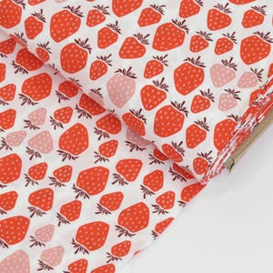 May include: White fabric with a repeating pattern of red strawberries and green leaves. Some strawberries are pink. The fabric is folded, revealing the pattern on the top layer and the edges of the fabric.