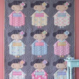 May include: A handmade quilt with nine doll figures in kimonos. The dolls have black hair and floral accents, set against a grey background with a floral pattern. The quilt is bordered with a gold trim. The kimonos are in various pastel colours.