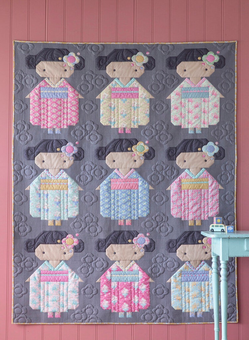 May include: A handmade quilt with nine doll figures in kimonos. The dolls have black hair and floral accents, set against a grey background with a floral pattern. The quilt is bordered with a gold trim. The kimonos are in various pastel colours.