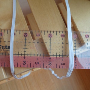 Puede incluir: Cord&oacute;n el&aacute;stico blanco medido con una regla. La regla muestra medidas en pulgadas y fracciones de pulgada. El cord&oacute;n el&aacute;stico mide aproximadamente 3 pulgadas de largo.
