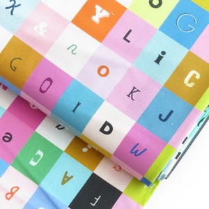 May include: Colorful fabric with a repeating pattern of alphabet letters in various colors. The letters are in different fonts and sizes, set against a grid of squares in shades of pink, blue, orange, and green. The fabric is folded over.