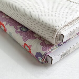 May include: Two rolls of fabric, one with a floral pattern in red, purple, and cream, and the other with a small dot pattern on a light background. The fabric is neatly rolled and stacked, with the edges visible. The fabric is from the "Tilda" collection.