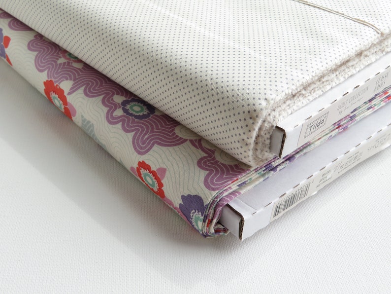 May include: Two rolls of fabric, one with a floral pattern in red, purple, and cream, and the other with a small dot pattern on a light background. The fabric is neatly rolled and stacked, with the edges visible. The fabric is from the "Tilda" collection.
