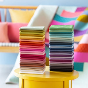 Op de afbeelding: Twee stapels kleurrijke stofstalen op een ronde gele tafel. De staal zijn gerangschikt in regenboogvolgorde, met de linker stapel beginnend met groen en de rechter stapel beginnend met blauw.