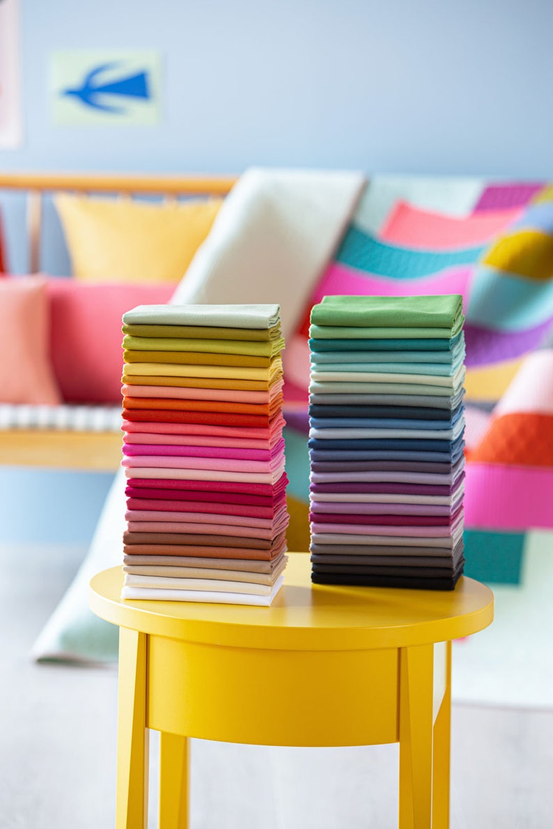 Op de afbeelding: Twee stapels kleurrijke stofstalen op een ronde gele tafel. De staal zijn gerangschikt in regenboogvolgorde, met de linker stapel beginnend met groen en de rechter stapel beginnend met blauw.