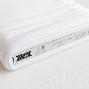 May include: A roll of white cotton fabric labeled "Tilda Solid Colors." The label indicates the fabric is 10 meters (10.93 yards) long. The fabric is a solid white color and appears to be tightly rolled.