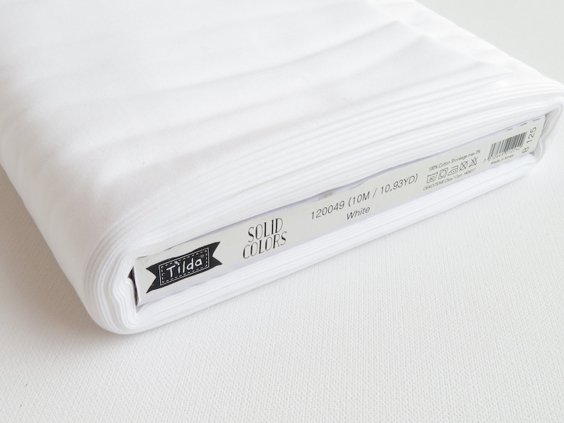 K&ouml;nnte beinhalten: Eine Rolle wei&szlig;en Baumwollstoffs mit der Aufschrift "Tilda Solid Colors". Das Etikett gibt an, dass der Stoff 10 Meter (10,93 Yards) lang ist. Der Stoff ist einfarbig wei&szlig; und scheint eng gerollt zu sein.