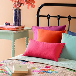 Op de afbeelding: Een bed met een kleurrijk patchwork-dekbed en een stapel kussens in verschillende tinten roze, oranje en blauw. Een boek ligt op het dekbed. Een vaas met een bloemstuk staat op een bijzettafel.