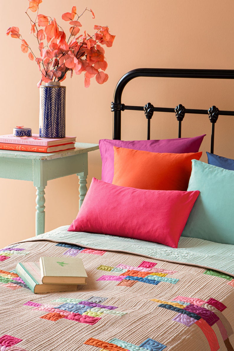 Op de afbeelding: Een bed met een kleurrijk patchwork-dekbed en een stapel kussens in verschillende tinten roze, oranje en blauw. Een boek ligt op het dekbed. Een vaas met een bloemstuk staat op een bijzettafel.