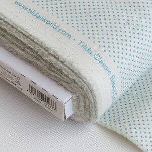 May include: A roll of white fabric with a pattern of small, evenly spaced turquoise dots. The fabric is labeled "Tilda Classic Basics Col" and "www.tildasworld.com". The edge of the fabric is frayed, and a white box is partially visible.