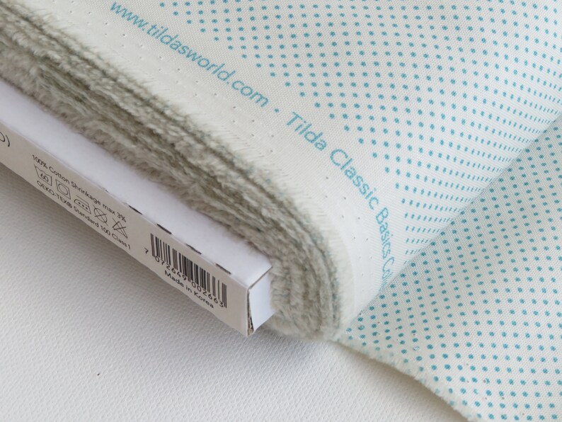 May include: A roll of white fabric with a pattern of small, evenly spaced turquoise dots. The fabric is labeled "Tilda Classic Basics Col" and "www.tildasworld.com". The edge of the fabric is frayed, and a white box is partially visible.