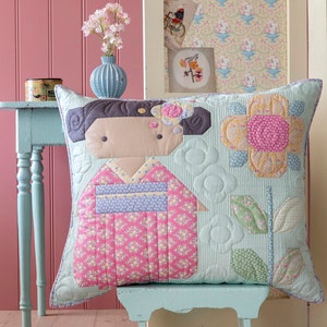 May include: A square cushion with a quilted geisha design in a pink kimono with floral patterns. The cushion has a light blue background with flower and leaf patterns. It sits on a small, light blue bench.