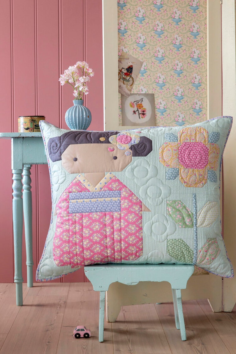 May include: A square cushion with a quilted geisha design in a pink kimono with floral patterns. The cushion has a light blue background with flower and leaf patterns. It sits on a small, light blue bench.