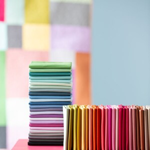 Op de afbeelding: Een stapel kleurrijke stofstalen op een roze tafel. De stalen zijn gerangschikt in een regenboogpatroon, met groen, blauw, paars, wit en zwart bovenaan en geel, oranje, rood, roze en bruin onderaan.