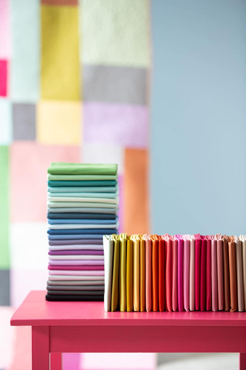 Op de afbeelding: Een stapel kleurrijke stofstalen op een roze tafel. De stalen zijn gerangschikt in een regenboogpatroon, met groen, blauw, paars, wit en zwart bovenaan en geel, oranje, rood, roze en bruin onderaan.