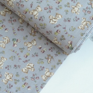 May include: A close-up of a light gray fabric with a repeating pattern of brown teddy bears, flowers, and toy blocks. The fabric is folded in half and the edge of the fabric is visible.