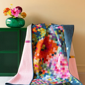 Op de afbeelding: Een kleurrijk quilt met een geometrisch patroon van vierkanten in tinten roze, oranje, geel, groen, blauw en paars. Het quilt is gevouwen en gedrapeerd over een groene kast.