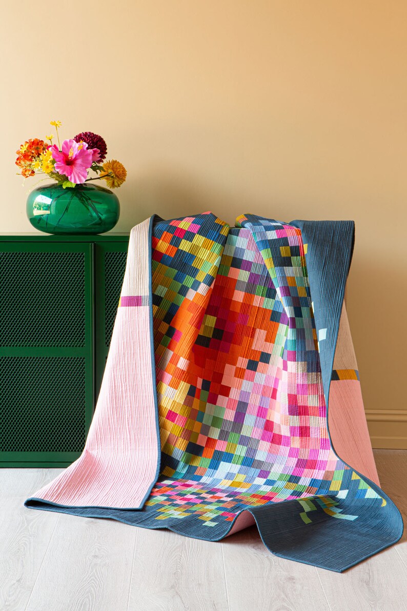 Op de afbeelding: Een kleurrijk quilt met een geometrisch patroon van vierkanten in tinten roze, oranje, geel, groen, blauw en paars. Het quilt is gevouwen en gedrapeerd over een groene kast.