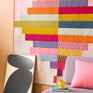 Op de afbeelding: Een kleurrijk, gestikt wandkleed met een geometrisch ontwerp in tinten roze, oranje, geel, groen, blauw en paars. Het wandkleed wordt getoond aan een muur naast een bank met een roze kussen en een geel theekopje.