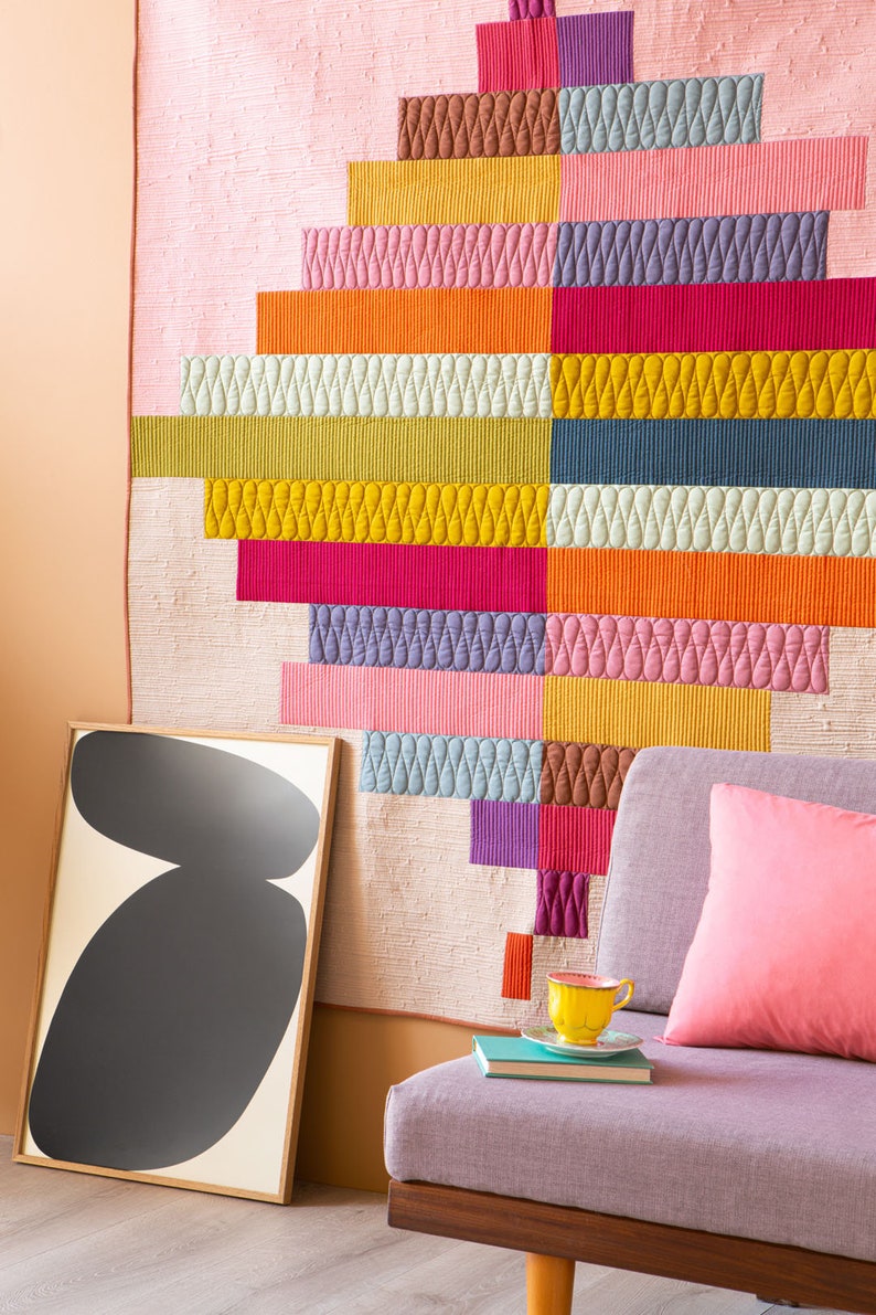 Op de afbeelding: Een kleurrijk, gestikt wandkleed met een geometrisch ontwerp in tinten roze, oranje, geel, groen, blauw en paars. Het wandkleed wordt getoond aan een muur naast een bank met een roze kussen en een geel theekopje.