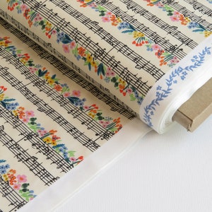 May include: A roll of fabric featuring musical notes and colorful floral accents. The fabric has a cream-colored background with black musical notation and rows of flowers in pink, yellow, and blue. The edge of the fabric has a blue floral border.