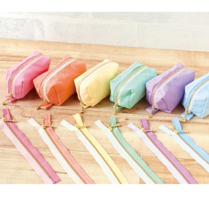 May include: A set of seven pastel-colored zippered pouches with gold zipper pulls. The pouches are arranged in a rainbow order from pink to light blue. The pouches are made of a soft, fabric material.
