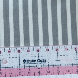 May include: A close-up of a white and gray striped fabric with a ruler measuring 6 inches. The ruler is pink and white with the words "Cute Cuts by Lori Holt of Bee in my Bonnet".