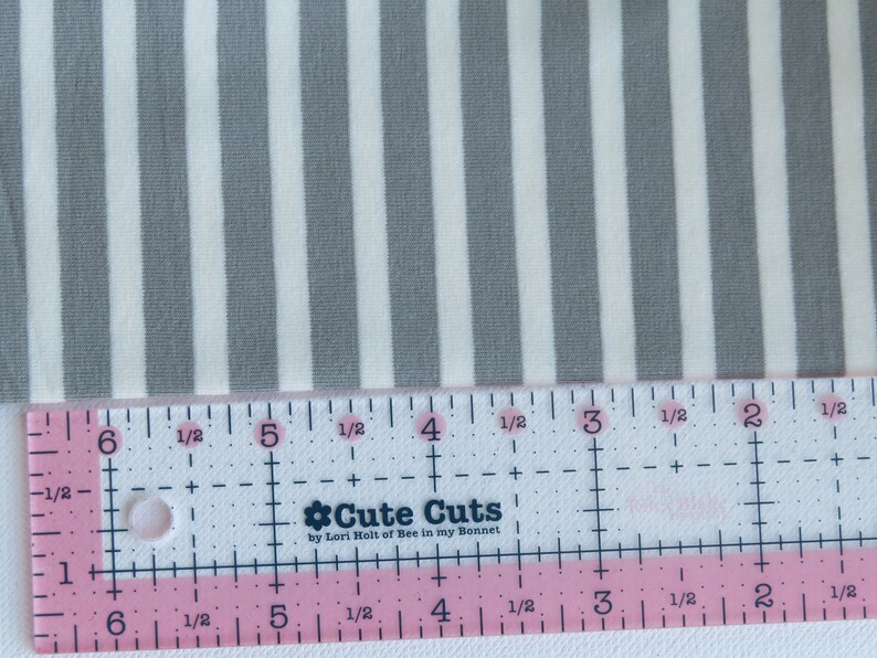 May include: A close-up of a white and gray striped fabric with a ruler measuring 6 inches. The ruler is pink and white with the words "Cute Cuts by Lori Holt of Bee in my Bonnet".