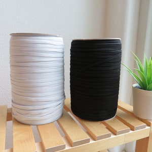 Puede incluir: Dos carretes de cord&oacute;n el&aacute;stico, uno blanco y otro negro, apilados en un estante de madera.