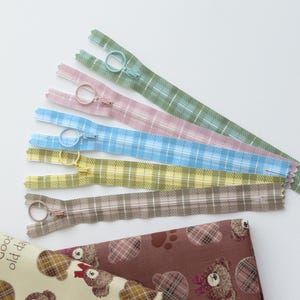 May include: A selection of patterned zippers in pastel shades of green, pink, blue, yellow, and brown. Each zipper has a circular pull. Coordinated fabric with teddy bear and plaid patterns is also visible.