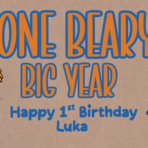Puede incluir: Una pancarta de cumpleaños con el texto "ONE BEARY BIG YEAR" en naranja y azul. Presenta un oso de dibujos animados con un gorro de fiesta y un ratón azul sosteniendo un globo. También se incluye el texto "Happy 1st Birthday Luka".