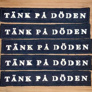 Könnte beinhalten: Fünf marineblaue rechteckige Aufnäher mit weißem Text "TÄNK PÅ DÖDEN". Die Aufnäher sind in einem ordentlichen Stapel angeordnet, wobei der Text auf jedem Aufnäher zentriert ist. Die Aufnäher bestehen aus einem strukturierten Material.