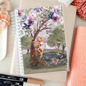 Pode incluir: Um caderno espiralado com uma ilustração de jardim de fadas. A capa apresenta uma pintura em aquarela de fadas, flores e um lago. O caderno está sobre uma superfície bege semelhante a couro, com marcadores e outros artigos de papelaria por perto.