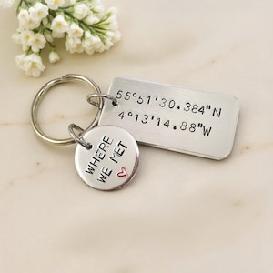 May include: A silver keychain with two charms. One charm is rectangular with coordinates, the other is round with the text "WHERE WE MET" and a small red heart. The keychain is on a marble surface.