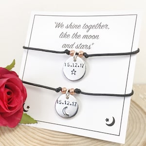 May include: Two black friendship bracelets with silver charms. The charms are round and have the date "15.12.17" engraved on them. One charm has a star and the other has a crescent moon. The card behind the bracelets reads "We shine together, like the moon and stars."