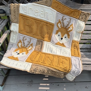 May include: A crocheted baby blanket with a patchwork design featuring a fox and deer motif in shades of brown, beige, and white.