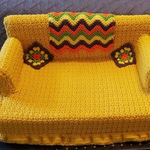 May include: A yellow crocheted couch with a brown, green, orange, and yellow chevron patterned throw pillow and two smaller floral patterned pillows.