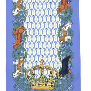 Könnte beinhalten: Hellblaues dekoratives Textil mit einem sich wiederholenden Muster aus Hunden, einer goldenen Krone und floralen Elementen. Das zentrale Design hat ein sich wiederholendes Muster aus grünen und weißen Blättern. Der Rand ist mit Hunden und floralen Designs verziert.