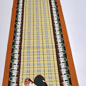 May include: A brown, green, and white table runner with a plaid pattern and a repeated image of a wirehaired fox terrier dog. The dog is in a walking pose with its tongue sticking out.