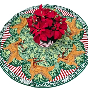 May include: A round Christmas tree skirt with a green floral pattern and illustrations of golden dogs with red ribbons. The border has red and white stripes. A pot of red poinsettias sits in the center.