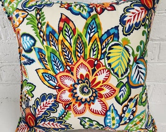 Pode incluir: Almofada decorativa quadrada com fundo creme e um padrão floral vibrante. O design apresenta grandes flores e folhas estilizadas em tons de vermelho, laranja, azul, verde e amarelo. A almofada tem uma borda contrastante.