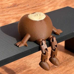 May include: A brown 3D-printed spherical figure with articulated limbs and a beige circular top section. The figure features a smooth, rounded body, jointed legs with defined feet, and outstretched arms with visible fingers, all rendered in a solid brown material.