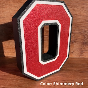May include: A three-dimensional letter O featuring a shimmery red glitter finish, bordered by a white outline and a black outer edge. The letter has a block-style shape with a hollow center and includes the text Color: Shimmery Red.