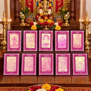 May include: A collection of ten rectangular prints featuring pink and white illustrations of Hindu deities, each framed in black with decorative borders. The prints are arranged in two rows on a wooden surface, with brass lamps on either side.