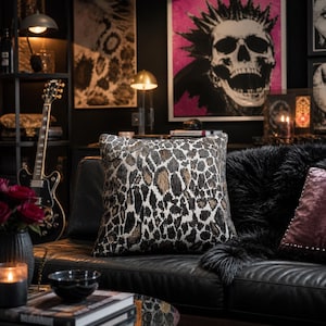 May include: Square decorative pillow with a brown, black, and white animal print pattern. The pillow is on a black leather couch. The room has a dark aesthetic with art and a guitar.