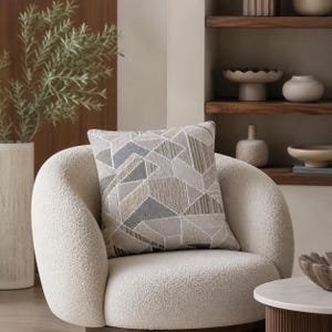 May include: A decorative throw pillow with a geometric pattern in shades of gray, beige, and blue rests on a cream-colored, curved armchair. The pillow's design features angular shapes and textured lines. The scene suggests a cozy living room setting.