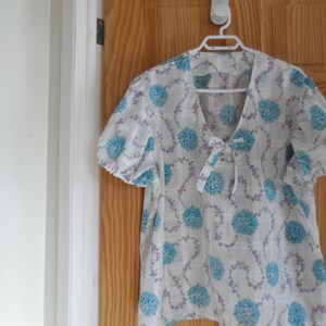 May include: A white blouse with a blue and purple floral pattern. The blouse has a V-neck and a bow at the neckline. The sleeves are puffed and end at the elbow.