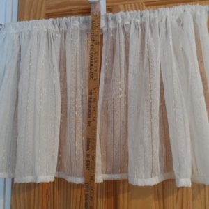 Puede incluir: Una cenefa blanca de voile con un patrón sutil, que mide aproximadamente 46 cm de largo. La cenefa está colgada de una barra de cortina blanca.