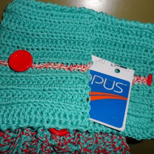 Op de afbeelding: Een turquoise gehaakte theedoek met een rode knoop en een witte en rode rand. De theedoek heeft een label met de tekst "OPUS".