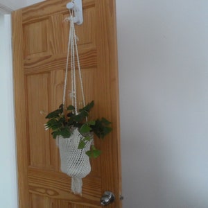 Peut inclure: Un porte-plante en macramé blanc avec une plante verte à l'intérieur suspendu à un crochet blanc sur une porte en bois.
